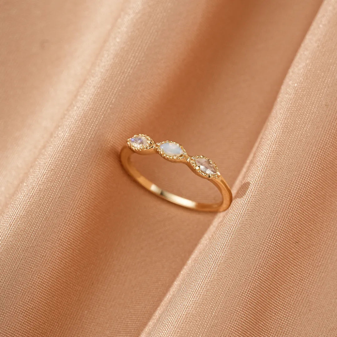 Bague en argent doré or sertie de trois pierres ovales, une opale blanche au centre et deux pierres de lune de chaque côté, sur un fond de satin couleur pêche.