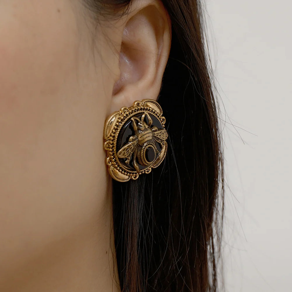 Boucles d'oreilles de fantaisie avec une abeille dorée en relief sur fond noir, ornées d'une pierre foncée, montées sur un support doré texturé.