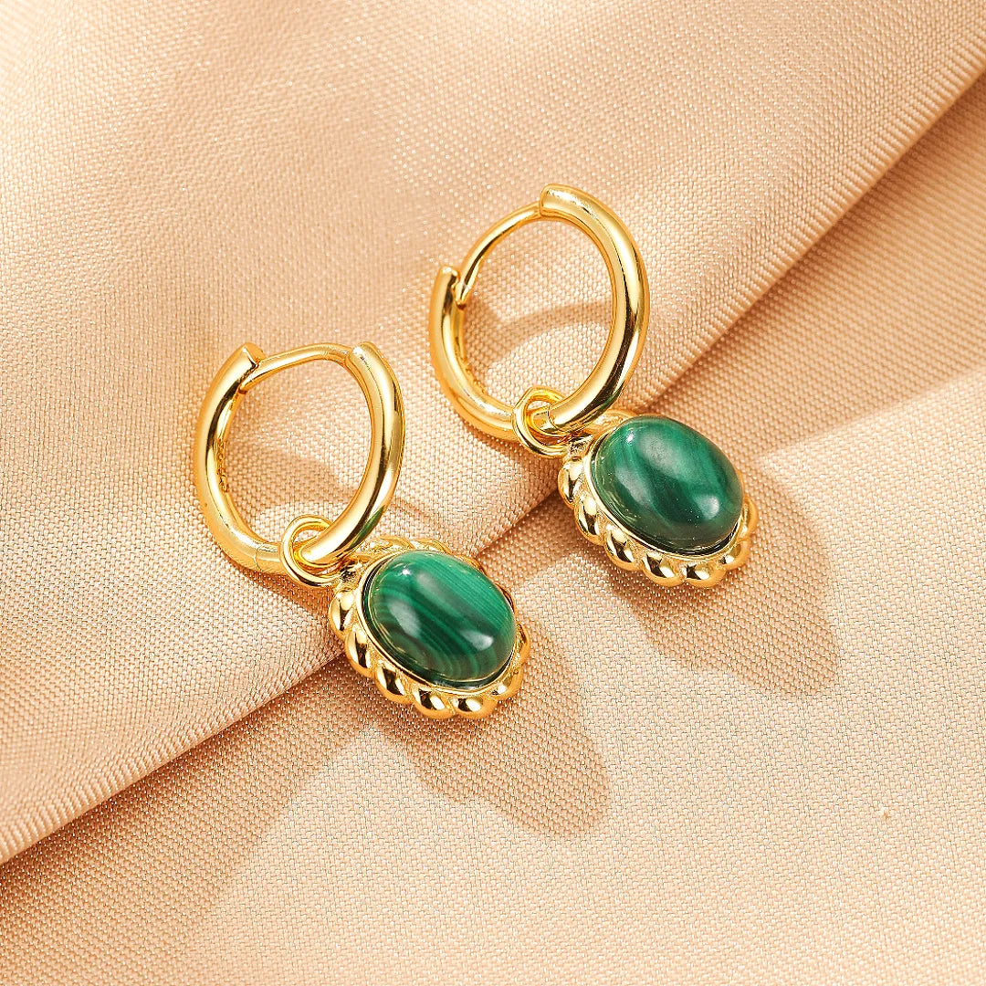 Boucles d'oreilles créoles dorées ornées de pierres ovales en malachite verte, montées sur un fond texturé beige.