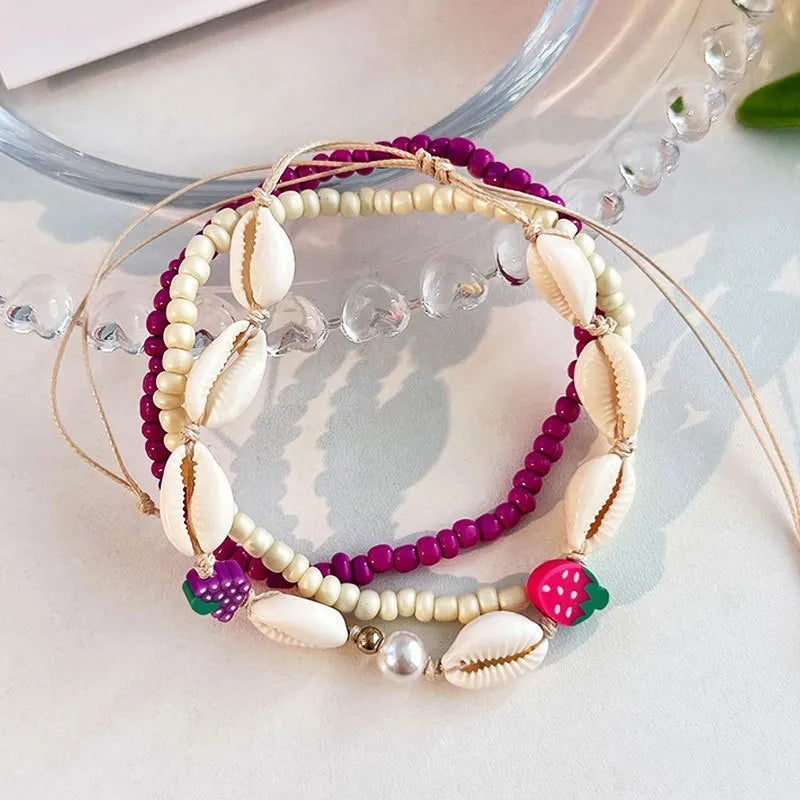 Ensemble de bracelets de cheville "Mer Profonde" avec des perles violettes, crème, des coquillages et des breloques en forme de fruits, dont une fraise et des raisins.