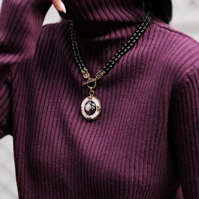 Gros plan sur un collier avec une double chaîne de perles noires et un pendentif rond orné d'une pierre blanche centrale entourée d'une finition dorée et noire, porté sur un pull col roulé prune.