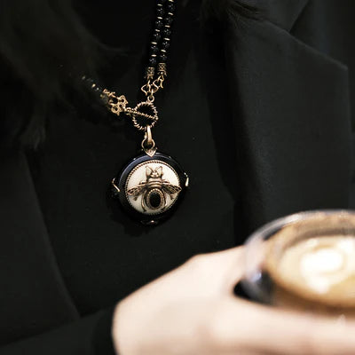 Collier "Reine Noire" avec un pendentif rond en agate noire orné d'une abeille dorée sur fond blanc, porté sur un fond noir.
