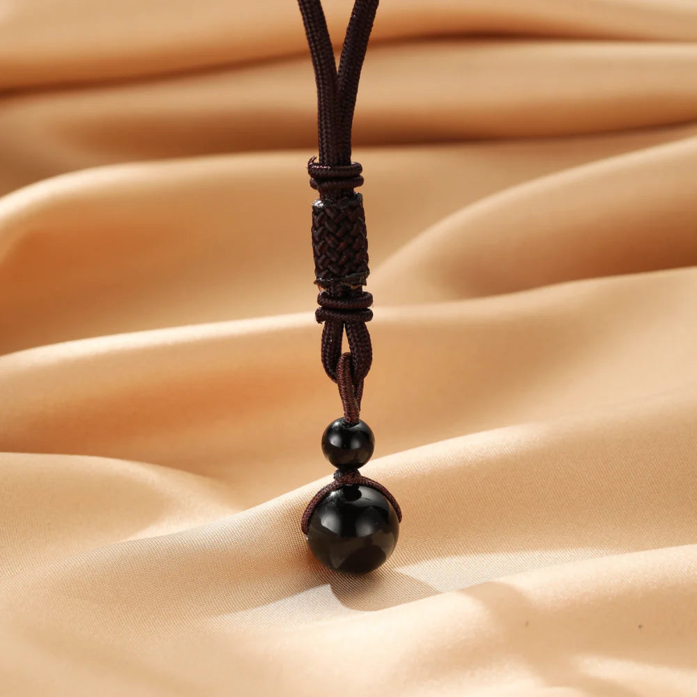 Collier avec une boule d'obsidienne polie et deux petites perles noires sur un cordon marron tressé, sur fond satiné beige.