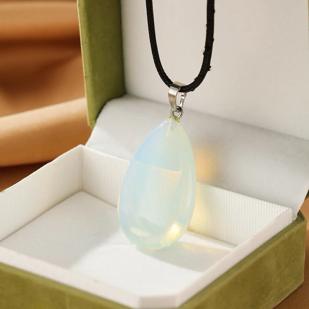 Un collier avec un pendentif en forme de goutte d'opale blanche laiteuse, monté sur un cordon noir, présenté dans une boîte cadeau verte.
