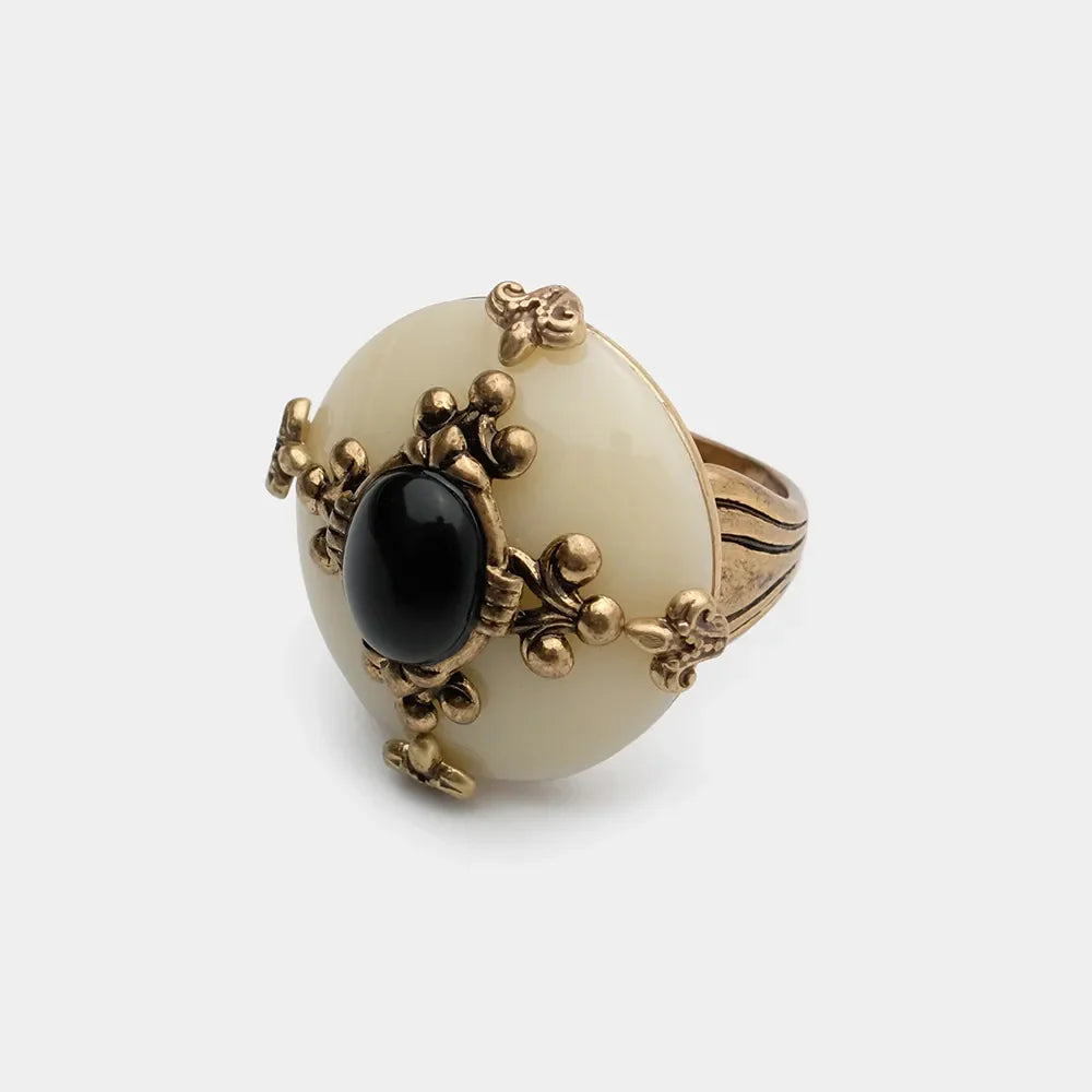 Bague "Pureté Mystique" avec une grande pierre d'agate noire ovale au centre, entourée de détails dorés ouvragés sur un fond blanc cassé, sur un fond gris clair.