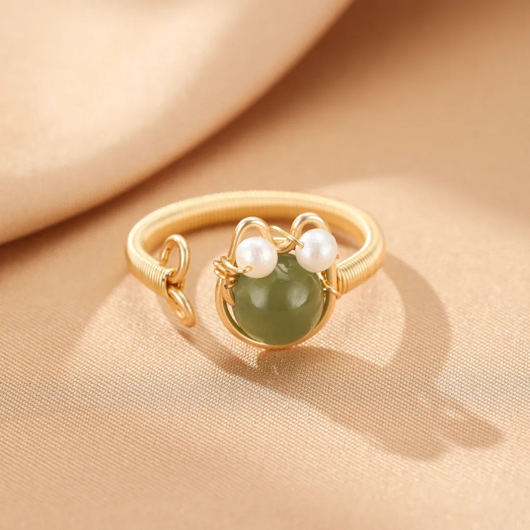 Bague ajusable dorée à l'or 14 carats ornée d'une pierre verte ronde et deux perles blanches formant un visage de chaton joyeux.