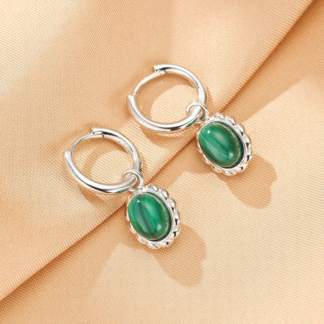Paire de boucles d'oreilles créoles en argent ornées de pierres de malachite ovales facettées, sur fond beige satiné.