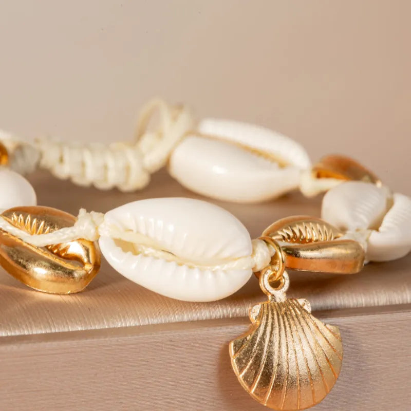 Gros plan d'un bracelet de cheville fait de coquillages blancs et de breloques dorées en forme de coquillage, sur un fond neutre.