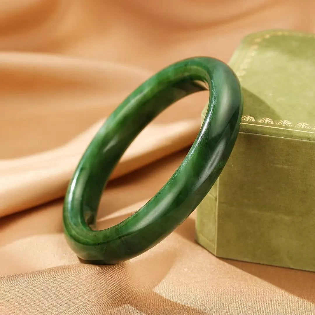 Bracelet jonc en jade "Verdure Éternelle" de couleur verte profonde, posé sur un tissu satiné beige à côté d'une boîte verte.