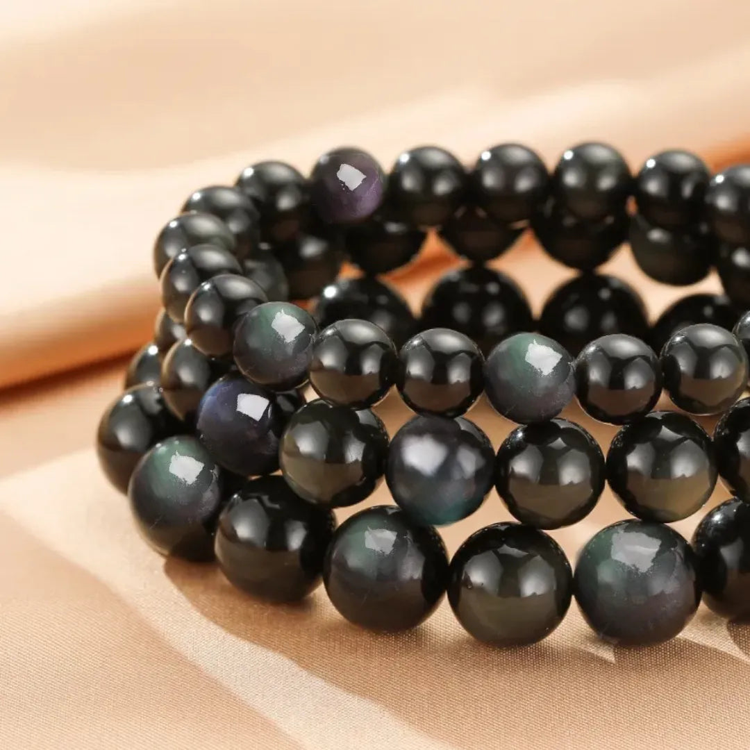 Bracelet fait de perles d'obsidienne arc-en-ciel noires, avec des reflets irisés de bleu et de violet visibles sous la lumière.