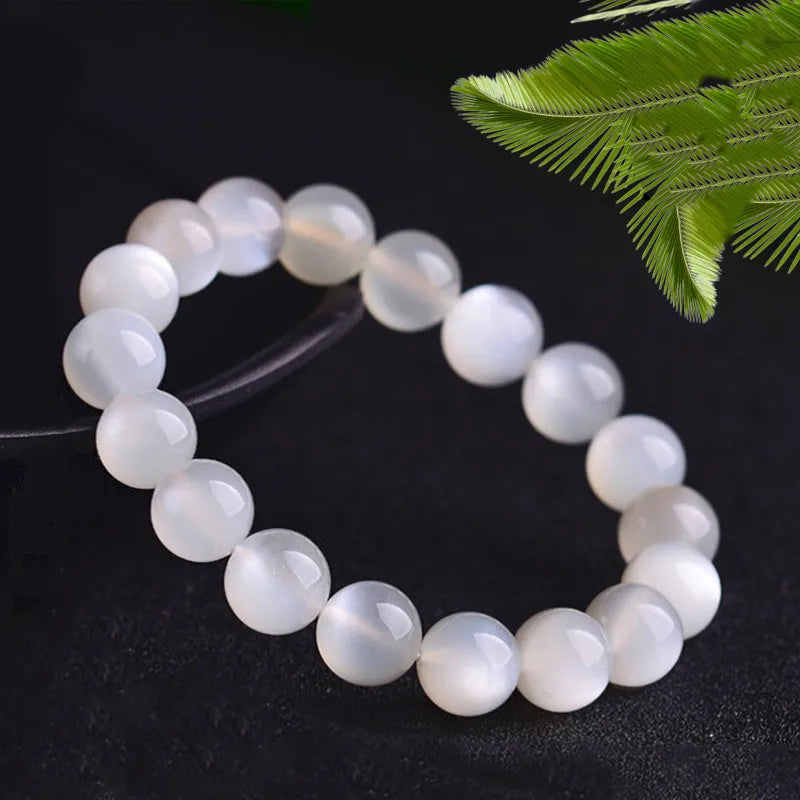 Bracelet "Élégance Lunaire" fait de perles rondes en pierre de lune blanche luminescente sur fond noir, avec une touche de verdure en arrière-plan.