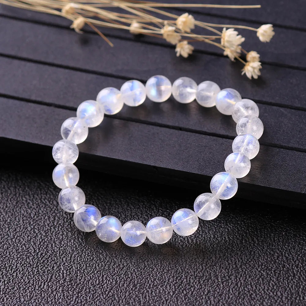 Bracelet "Rêve Doux" en pierre de lune, composé de perles rondes translucides aux reflets bleutés et irisés, posé sur un fond sombre avec des herbes séchées en arrière-plan.