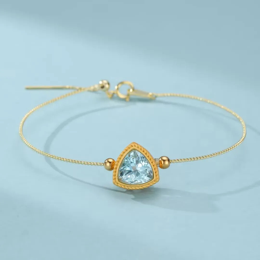 Bracelet fin en argent doré avec une topaze bleue en forme de triangle taillée, rehaussé de deux petites perles dorées de chaque côté du pendentif.