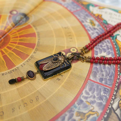 Collier avec pendentif rectangle noir orné d'une cigale dorée et d'une pierre rouge, sur un fond illustré de cartes célestes et d'un tour de cou en perles rouges.