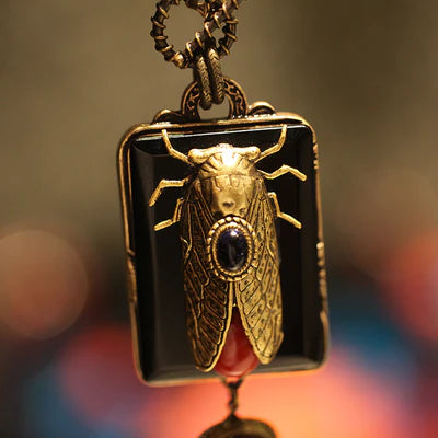Collier fantaisie "Beauté Naturelle" avec un pendentif rectangulaire noir orné d'une cigale dorée incrustée d'une pierre bleue et d'une goutte rouge.
