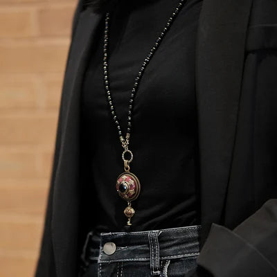 Collier Agate "Merveille d'Antan" avec pendentif rond orné de détails dorés et d'une pierre sombre, porté sur un haut noir et une veste.