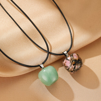 Deux colliers avec des pendentifs en forme de cœur, l'un en aventurine verte et l'autre en rhodonite rose, sur un fond satiné beige.