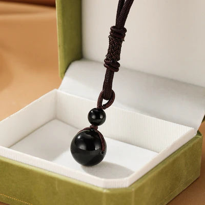Collier avec une boule d'obsidienne noire lisse et une plus petite sur un cordon marron tressé, présenté dans une boîte cadeau verte.