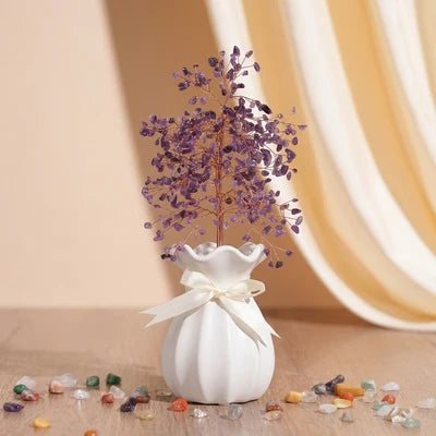 Arbre de Vie fait de pierres d'améthyste violettes et d'un fil de cuivre, placé dans un pot blanc orné d'un ruban, entouré de petites pierres polies.